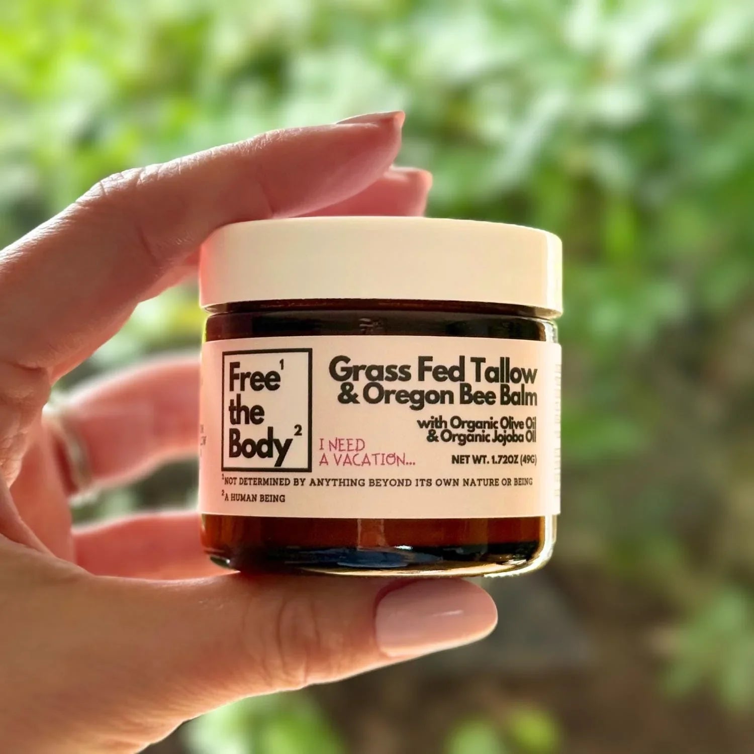 Hand holding a jar of 'Free the Body' Grass Fed Tallow & Oregon Bee Balm against a blurred green background