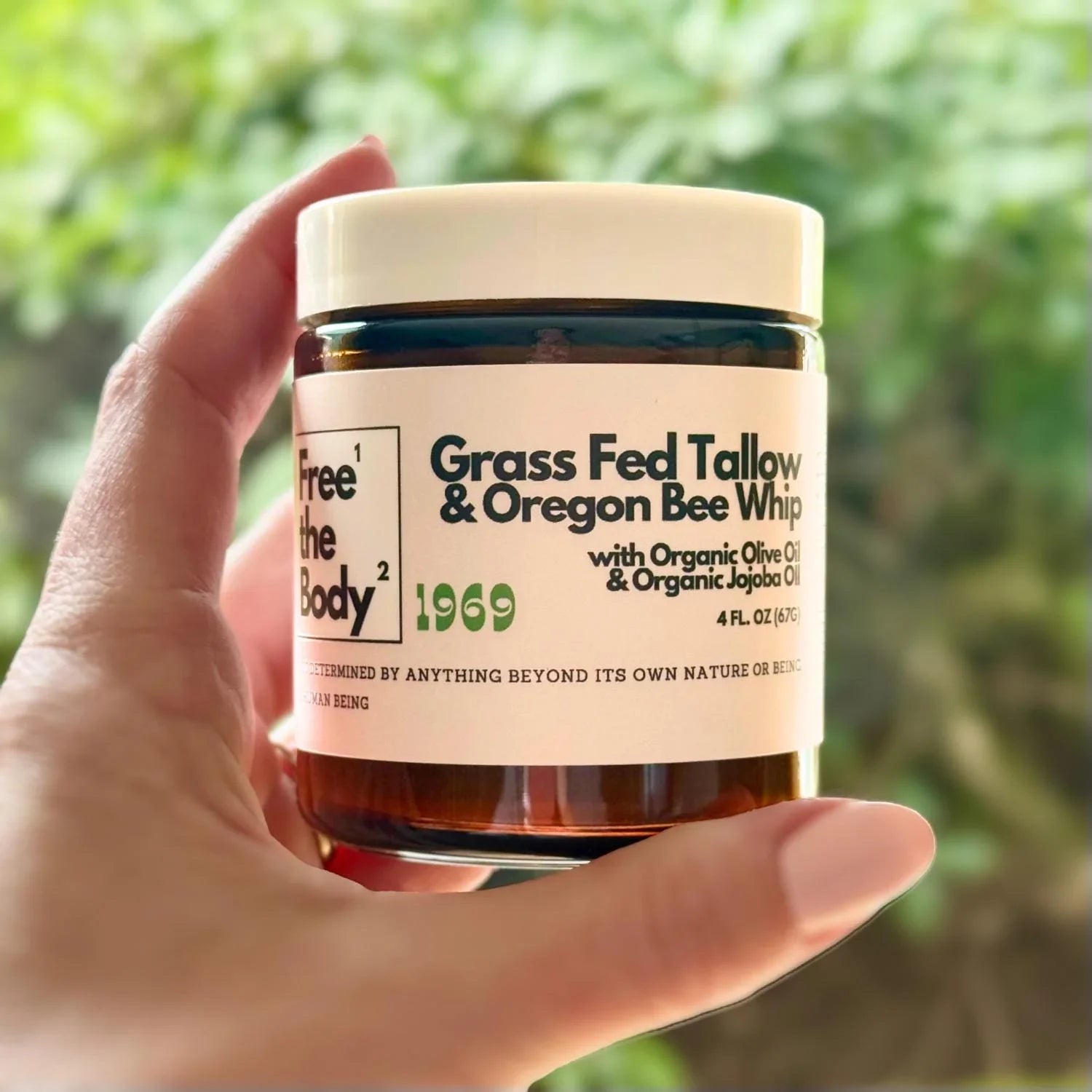 Hand holding a jar of 'Free the Body' Grass Fed Tallow & Oregon Bee Whip against a blurred green background