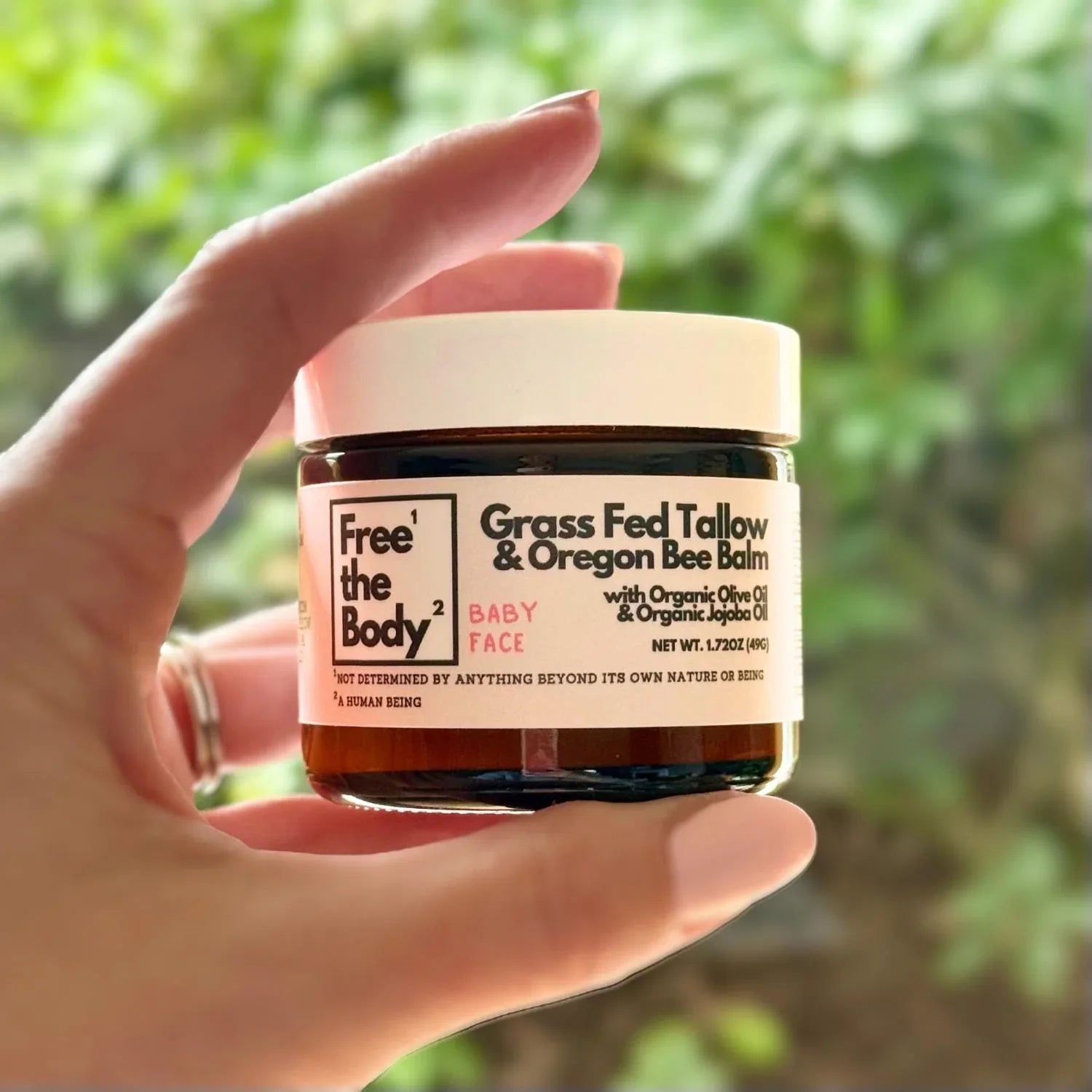 Hand holding a jar of 'Free the Body' Grass Fed Tallow balm against a blurred green background