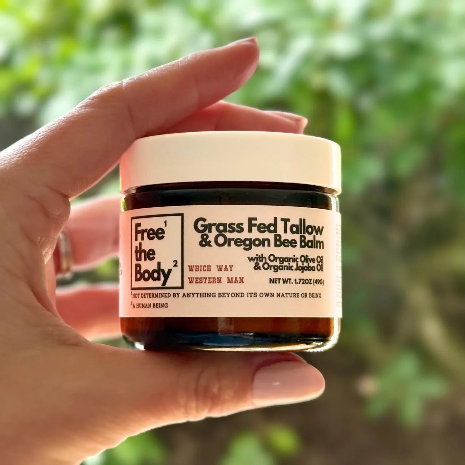 Hand holding a jar of 'Free the Body' Grass Fed Tallow & Oregon Bee Balm against a blurred green background