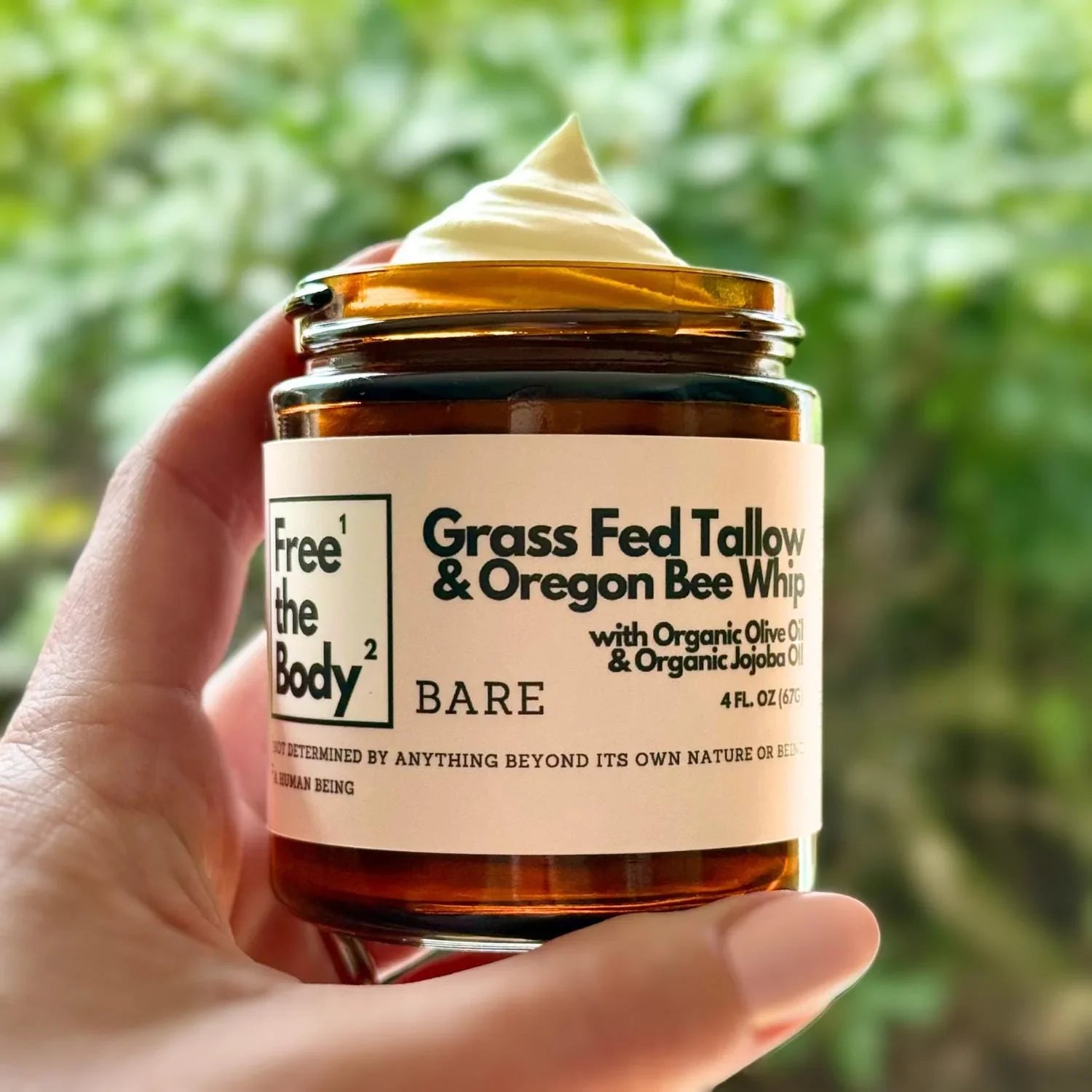 Hand holding a jar of 'Free the Body' Grass Fed Tallow & Oregon Bee Whipped Tallow Balm with a blurred green background