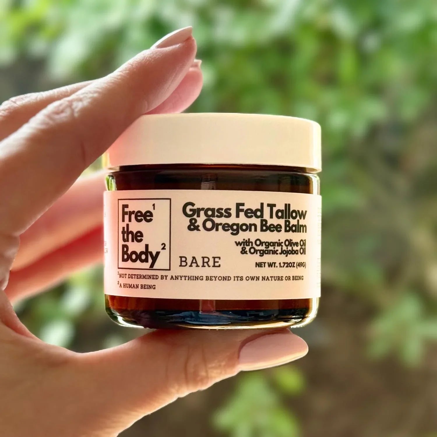 Hand holding a jar of 'Free the Body' Grass Fed Tallow & Oregon Bee Balm against a blurred green background